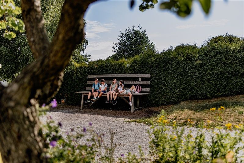 Vier Kinder sitzen auf einer übergroßen Bank in einem Garten, umgeben von Bäumen und Sträuchern.