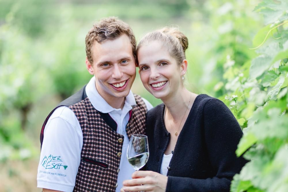 Ein lächelndes Paar in einem Weinberg, der Mann trägt eine Weste, die Frau hält ein Weinglas.