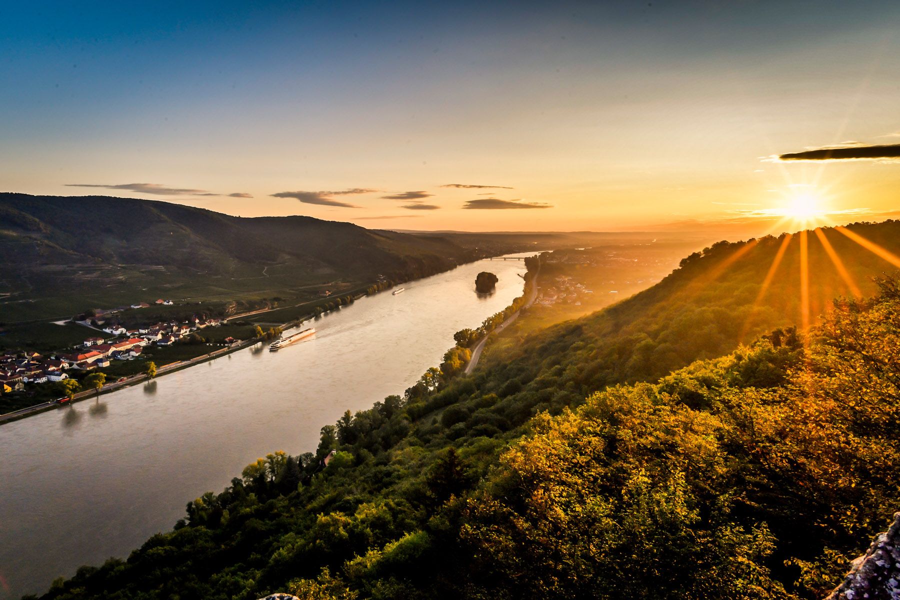 Sonnenuntergang über einem Fluss mit Hügeln und Dörfern im Hintergrund.
