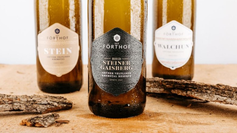 Drei Weinflaschen der Marke F&ouml;rthof auf einem Holztisch mit Rindenst&uuml;cken.