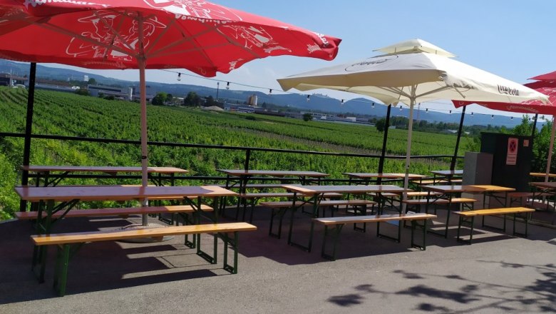 Biergarten mit Tischen und Sonnenschirmen vor einem Weinberg.