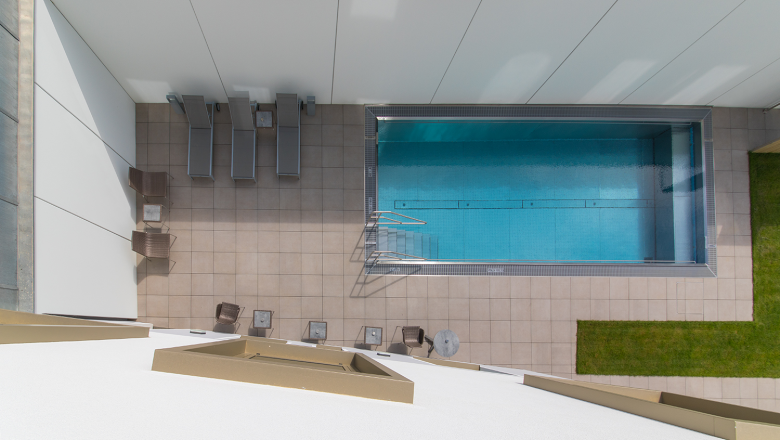 Draufsicht auf einen rechteckigen Pool mit Liegest&uuml;hlen und Tischen auf einer gefliesten Terrasse.