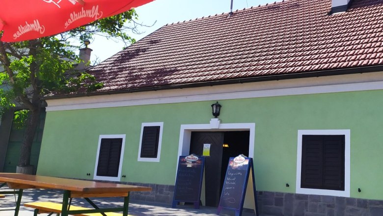 Exterior view of a traditional wine tavern with green paint and red roof tiles. There are wooden benches and a red parasol in front of the building.