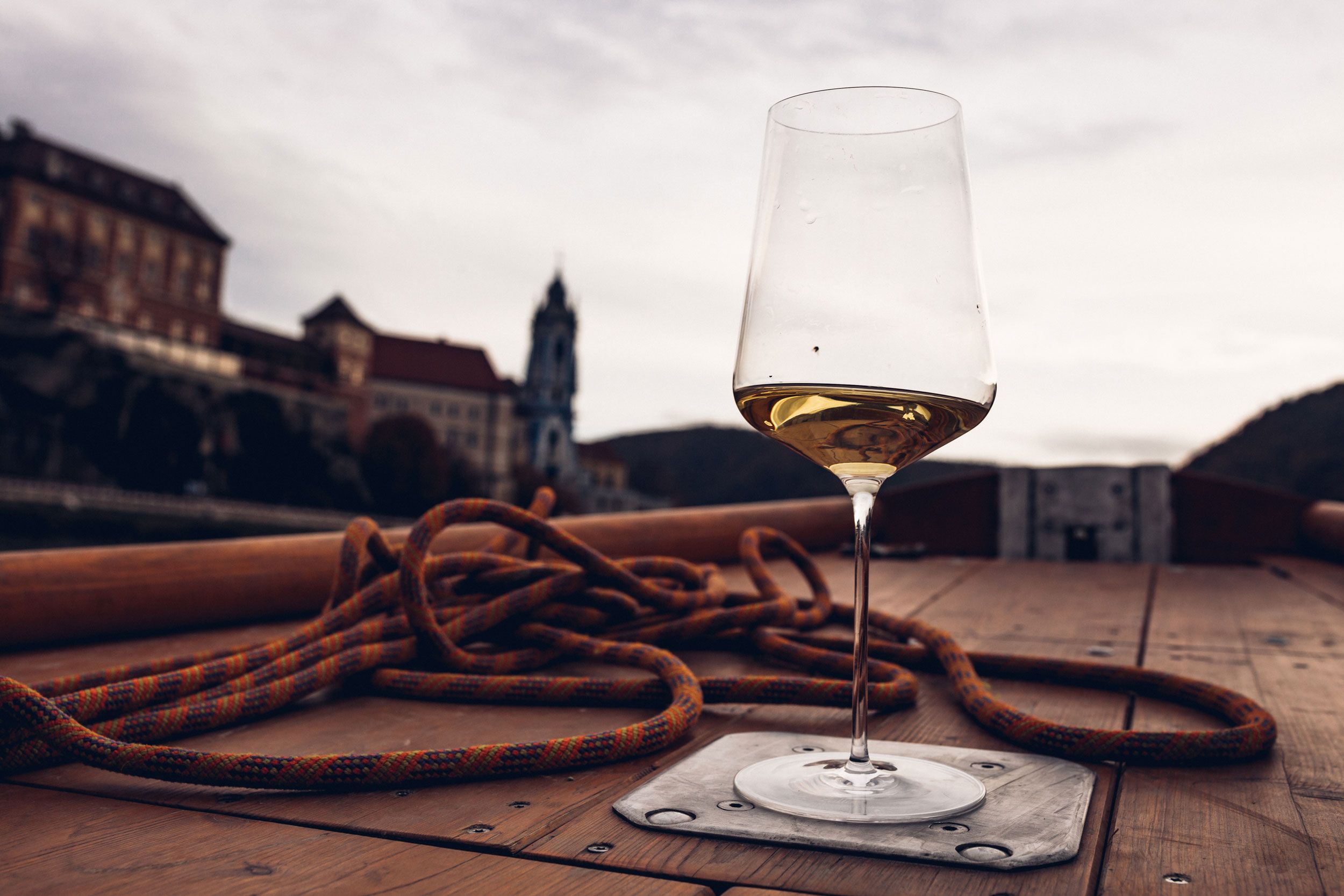 Ein Weinglas mit Weißwein steht auf einem Boot, im Hintergrund ist die Kirche von Dürnstein unscharf zu sehen.