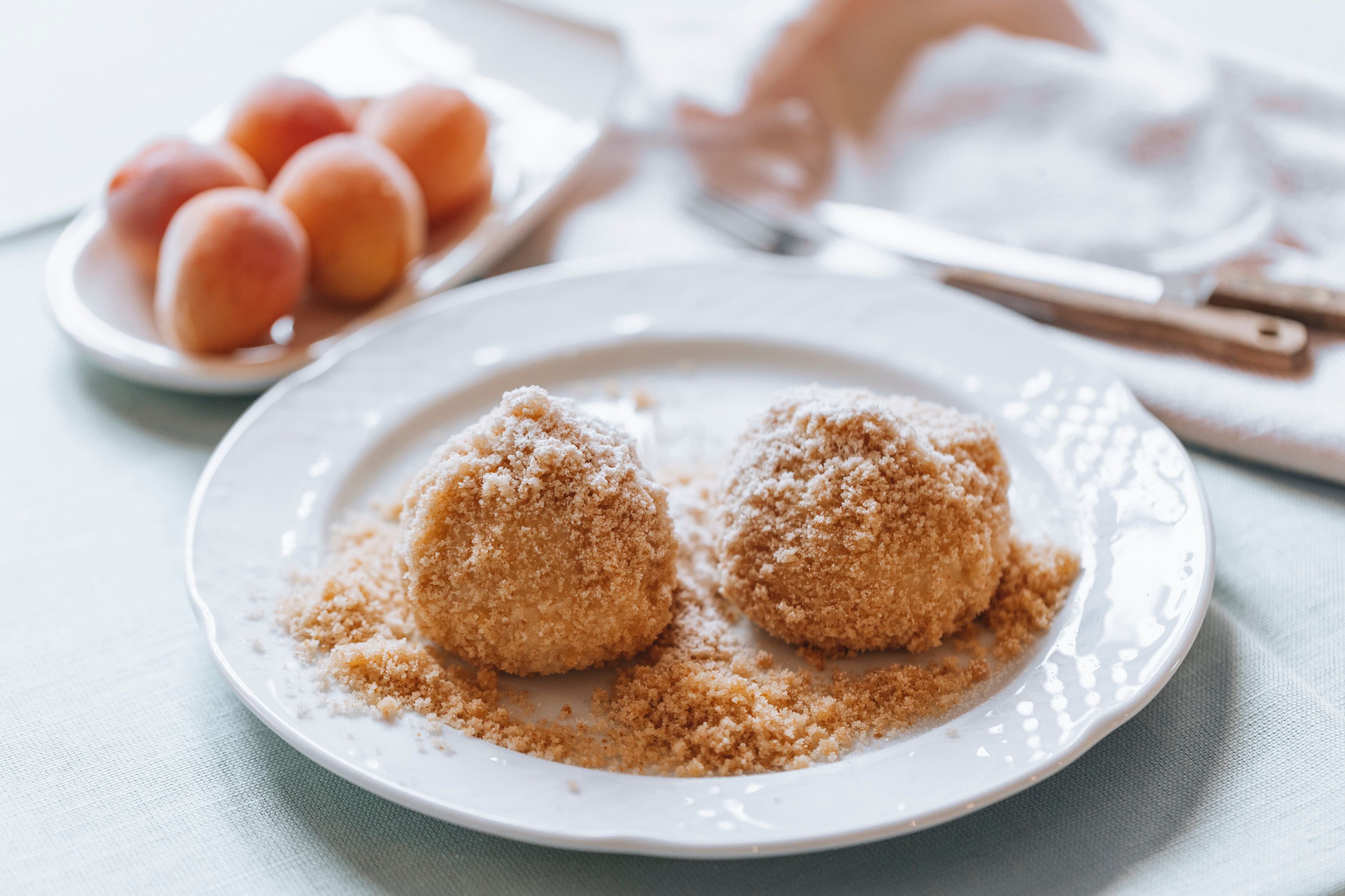 Zwei Marillenknödel auf einem Teller mit Bröseln, im Hintergrund Aprikosen.