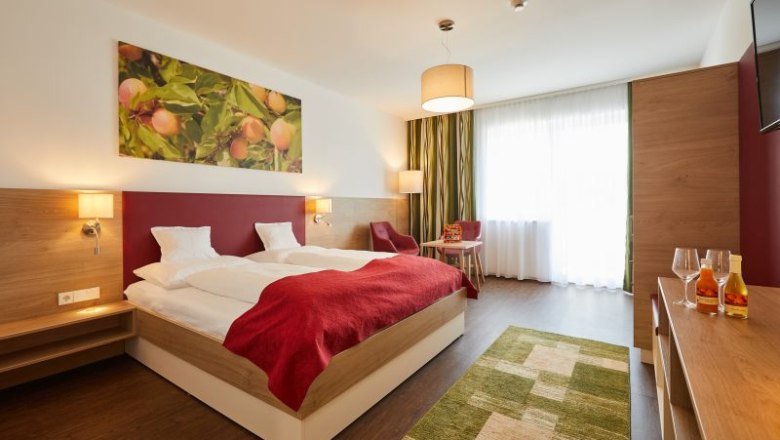 Modern eingerichtetes Hotelzimmer mit Doppelbett, roter Decke, Holzm&ouml;beln und gro&szlig;em Fenster. An der Wand h&auml;ngt ein Bild mit Fr&uuml;chten.