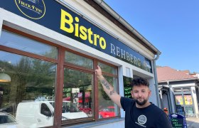 Ein Mann zeigt auf ein Schild mit der Aufschrift 'Bistro Rehberg'.