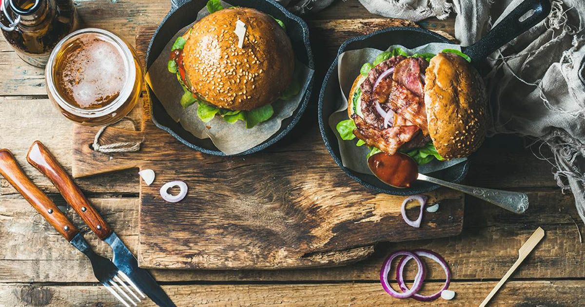 Zwei Burger in Pfannen mit Bier und Besteck auf Holztisch.