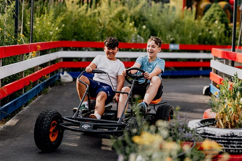 Zwei Jungen fahren lachend in einem Go-Kart auf einer Strecke mit rot-weißen Zäunen.