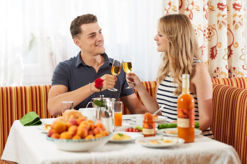 Ein Paar stößt mit Sektgläsern beim Frühstück an, umgeben von Obst und Getränken auf einem gedeckten Tisch.
