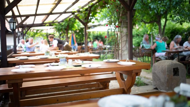 A guest garden with wooden tables and benches, surrounded by green plants. People sit in the background and enjoy their meals.