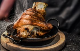 Knusprige Schweinshaxe in einer Pfanne auf einem Holzbrett, mit Salz bestreut.