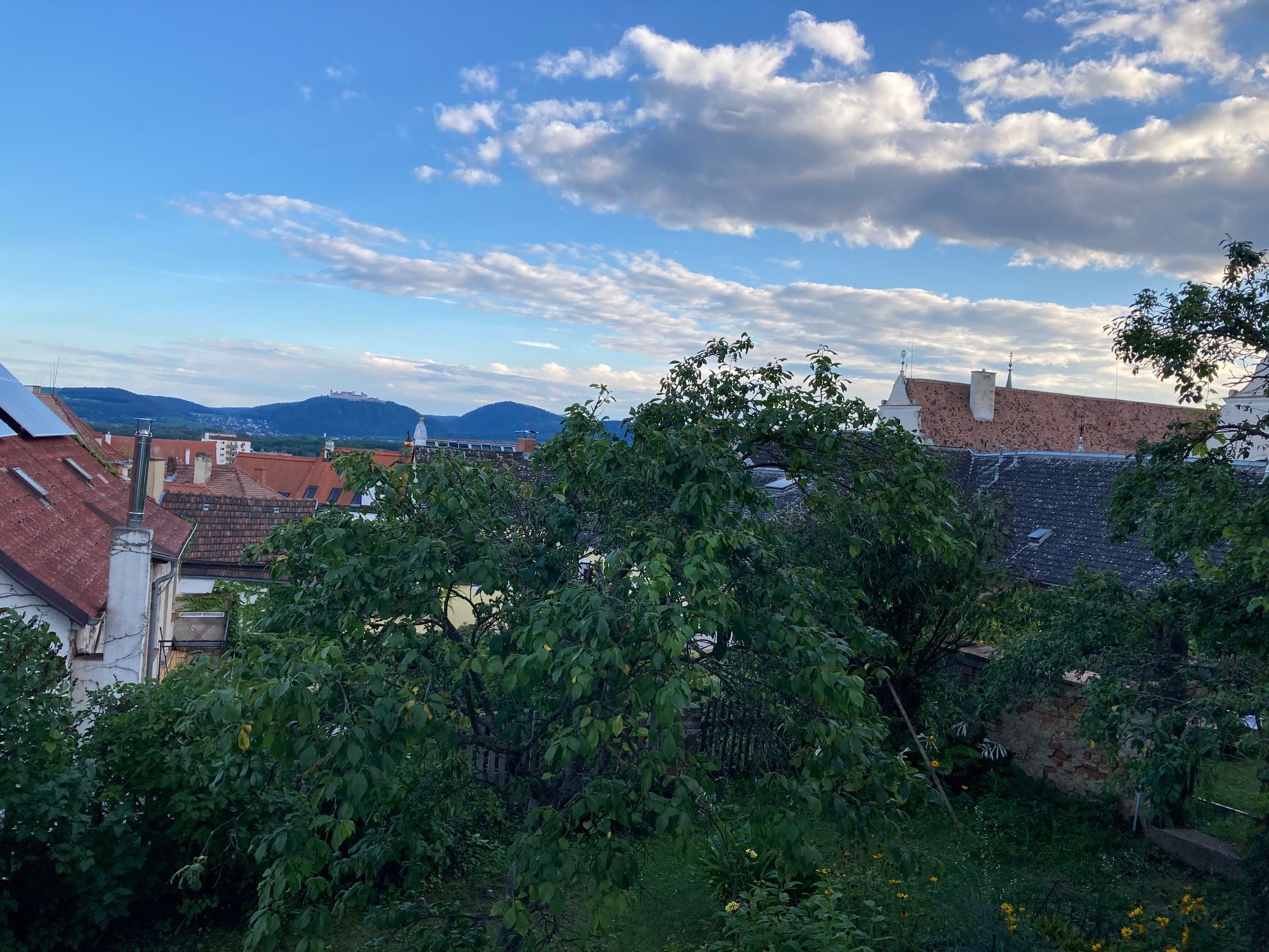 Blick über Dächer und Bäume auf Hügel und blauen Himmel mit Wolken.