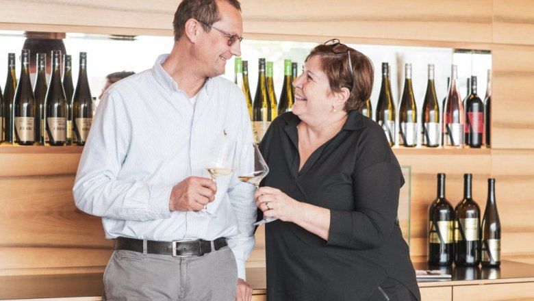 Ein Mann und eine Frau stoßen mit Weingläsern an, im Hintergrund Weinflaschen.