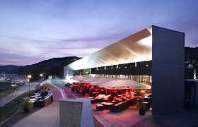 Modernes Geb&auml;ude am Donau-Ufer bei D&auml;mmerung mit beleuchteter Terrasse und Sitzbereichen.