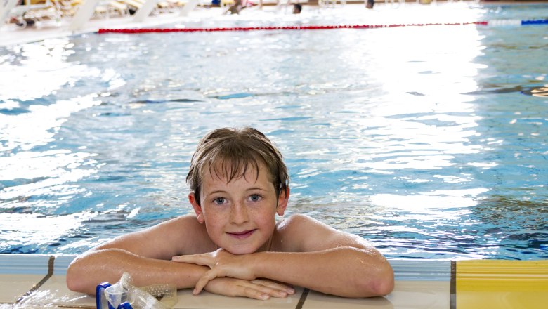 A boy is leaning against the edge of an indoor pool, smiling and wearing swimming goggles.