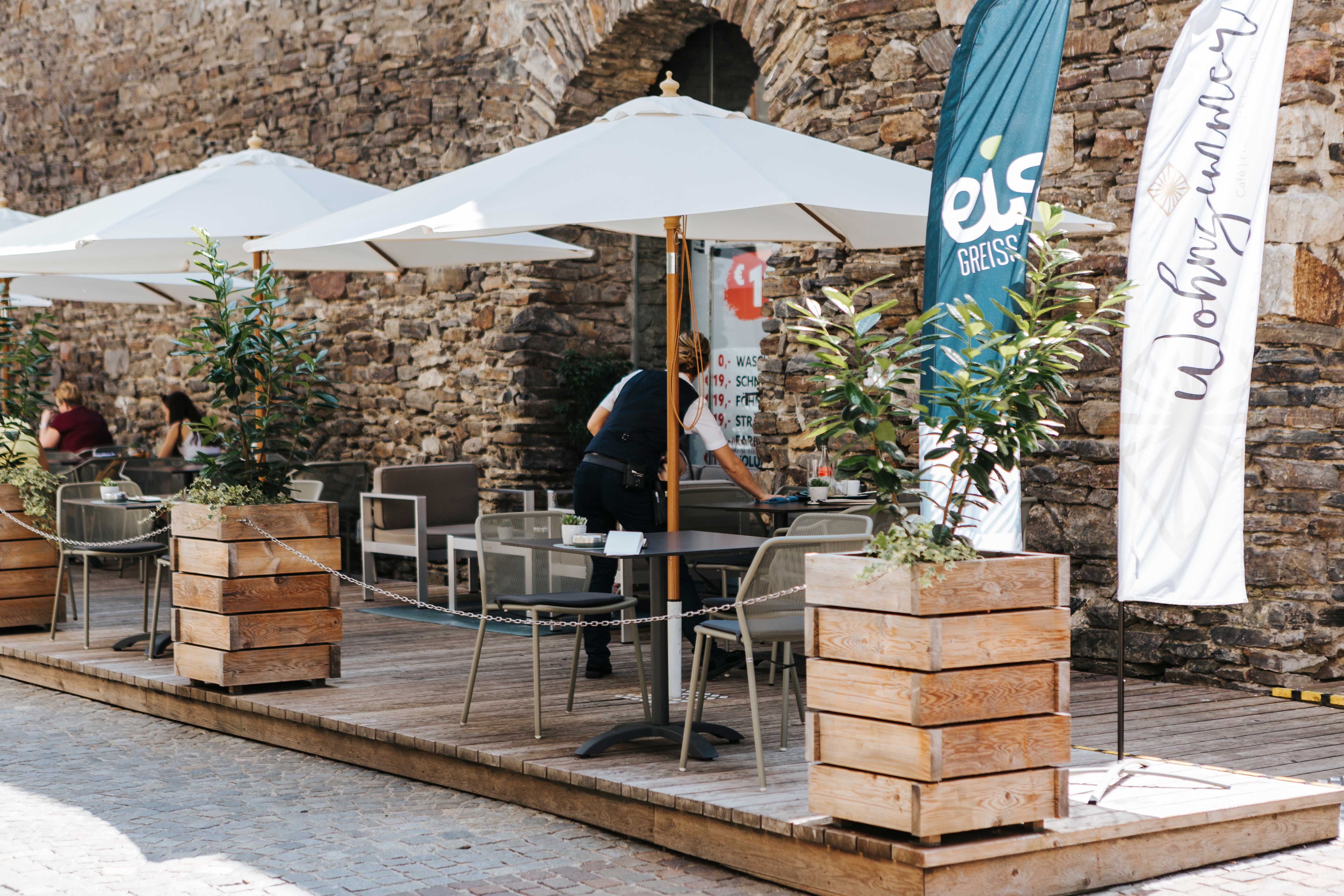 Außenbereich eines Cafés mit Sonnenschirmen und Holzkisten als Pflanzgefäße vor einer Steinmauer.