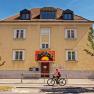 Yellow building with red entrance and graffiti sign. A cyclist rides past. Blue sky and trees in the background.