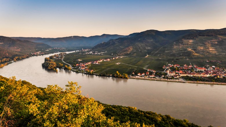 Panoramic view of a river, surrounded by hills and villages, at sunset.