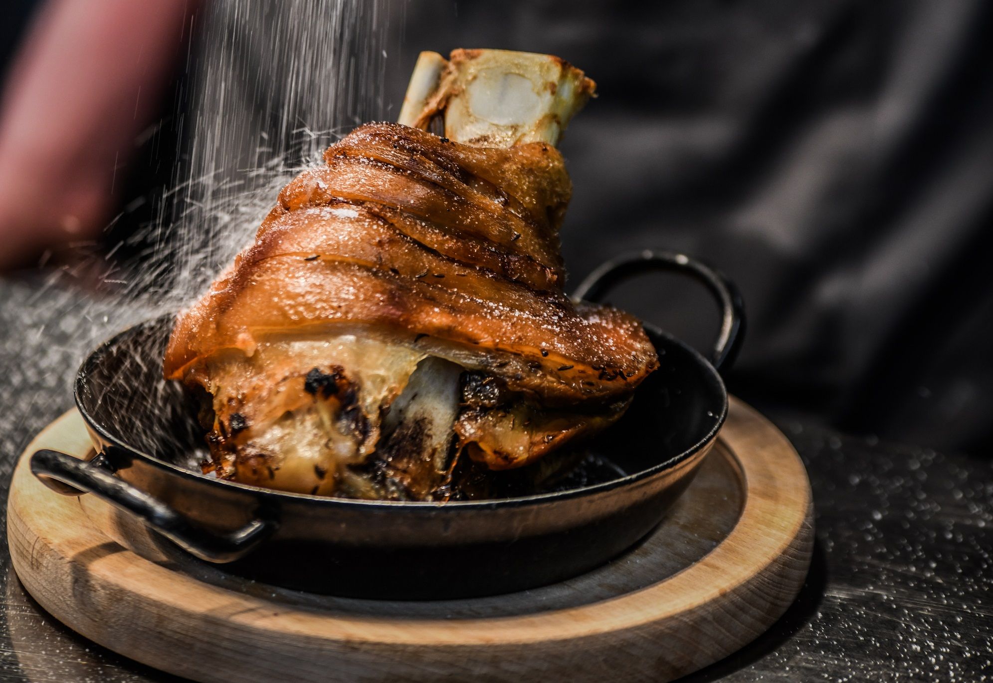 Knusprige Schweinshaxe in einer Pfanne auf einem Holzbrett, mit Salz bestreut.