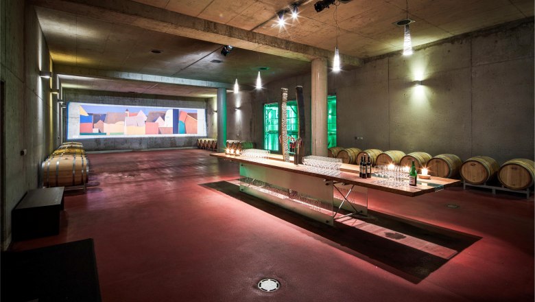 Ein moderner Weinkeller mit Holzfässern und einem langen Tisch mit Gläsern und Flaschen.