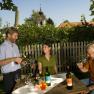 Three people enjoy wine in the garden, with a wooden fence and a church in the background.