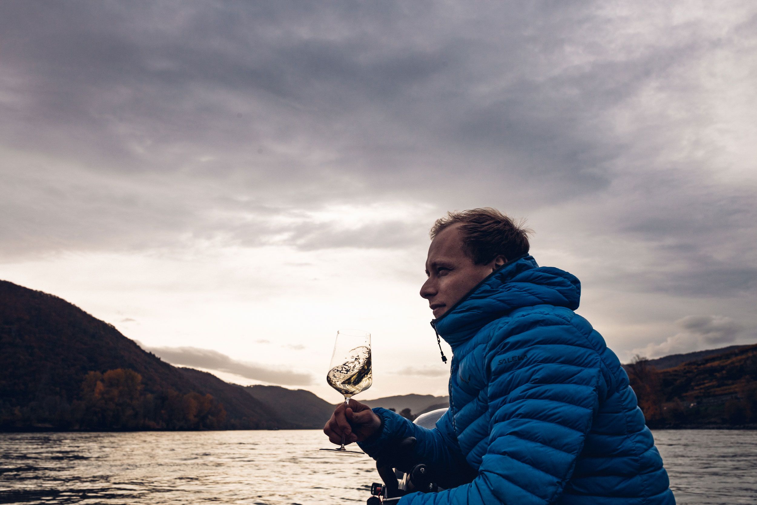 Person in blauer Jacke hält ein Weinglas auf einem Boot vor einer Flusslandschaft.