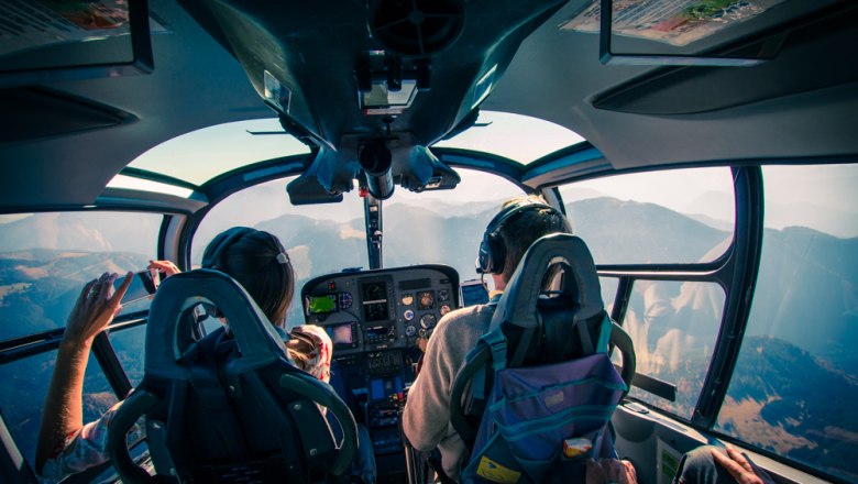 View from the cockpit of a helicopter with two people flying over a mountain landscape.