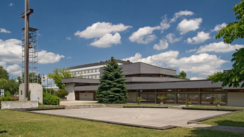 Modern church with bell tower and cross, surrounded by green spaces and trees, under a blue sky with clouds.