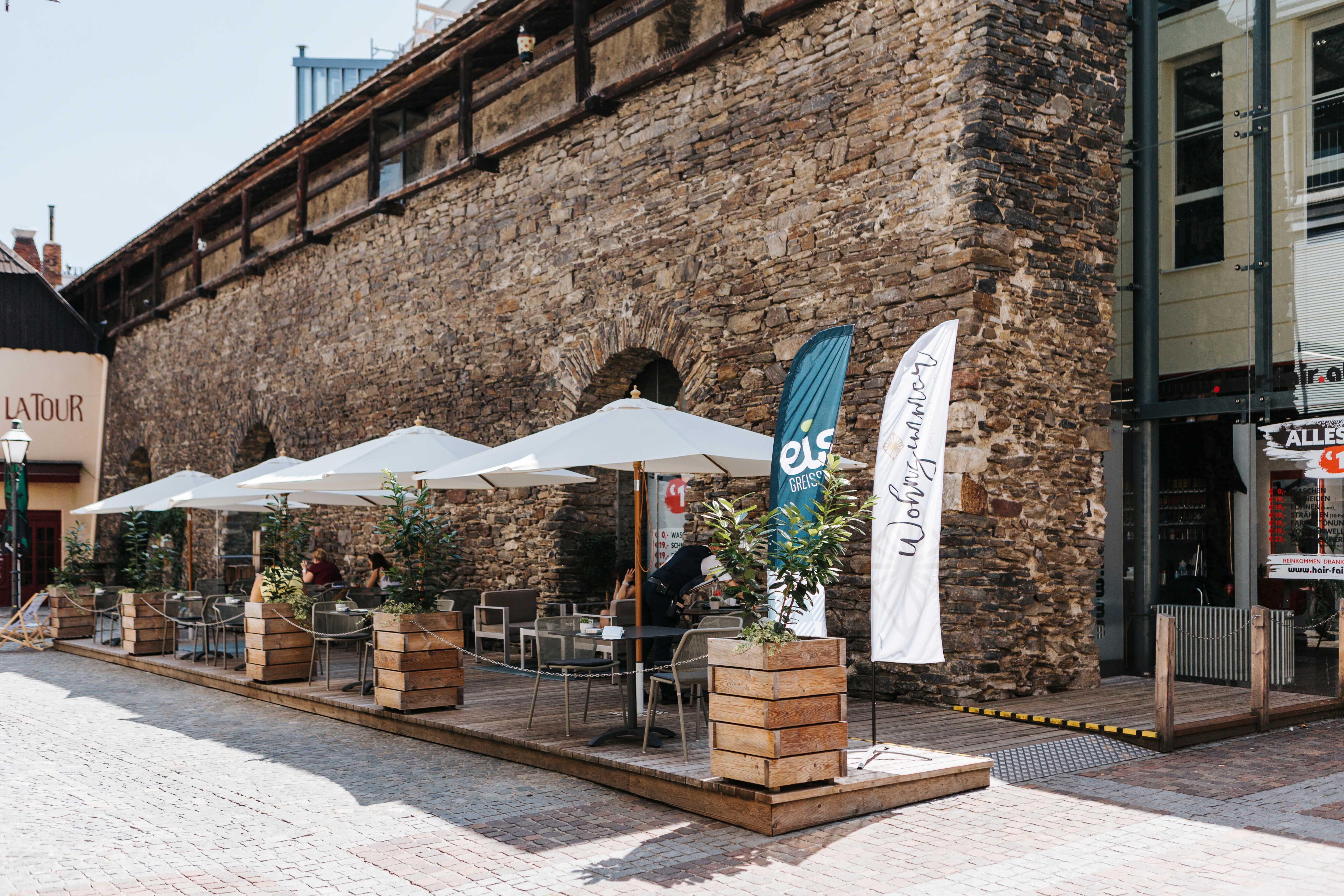 Außenterrasse eines Cafés mit Sonnenschirmen und Pflanzen vor einer alten Steinmauer.
