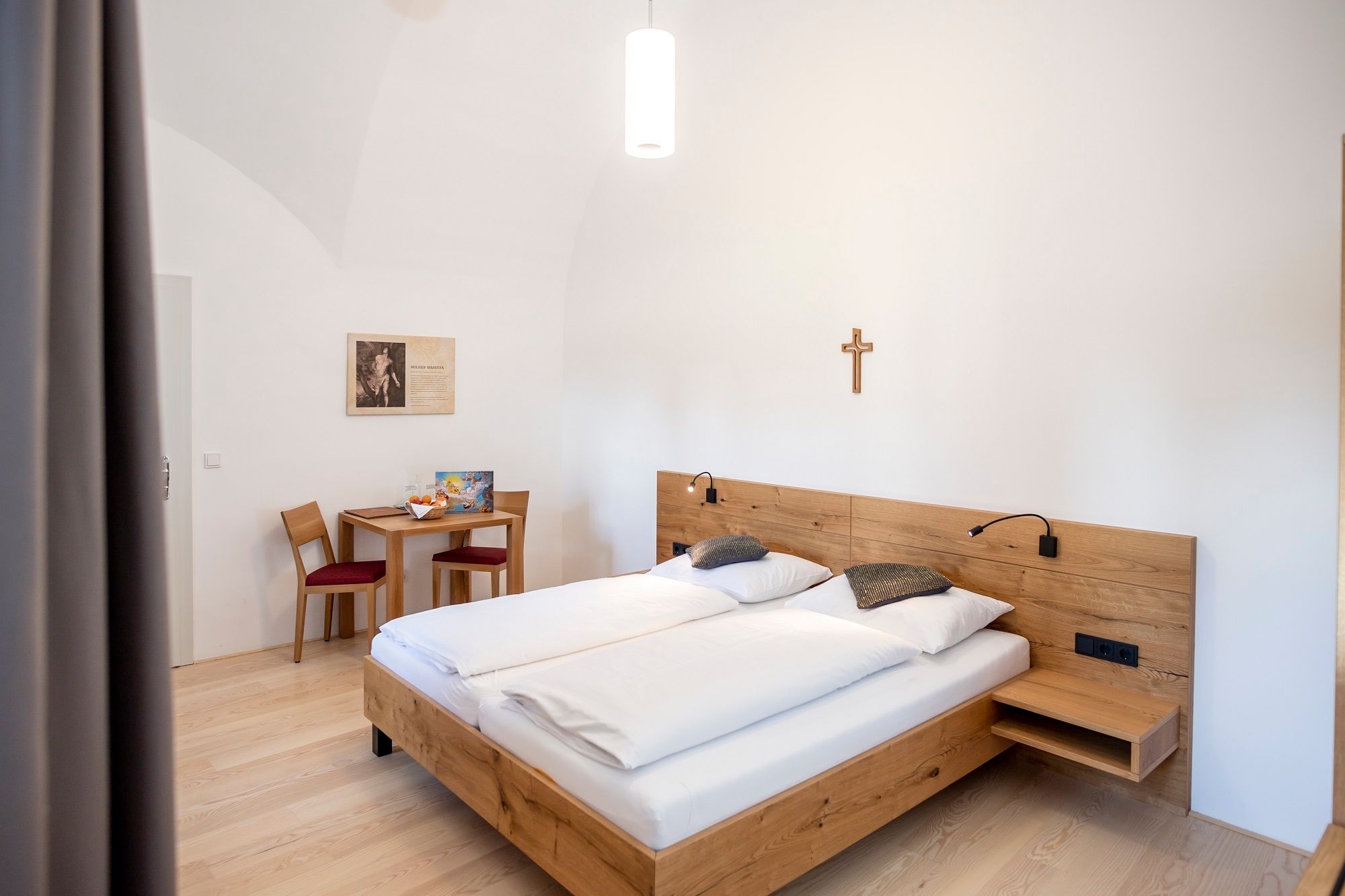 Ein schlicht eingerichtetes Gästezimmer mit Doppelbett, Holzmöbeln und einem Kreuz an der Wand.