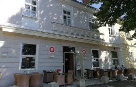 Außenansicht eines Cafés mit Terrasse und Sitzgelegenheiten unter einem Baum.