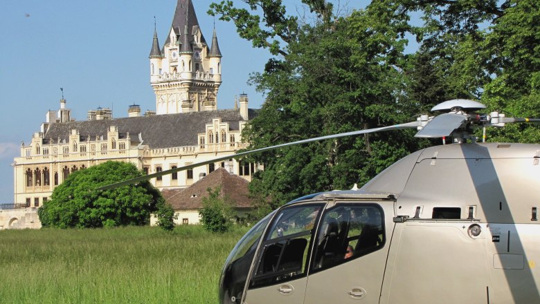 A helicopter stands on a meadow in front of a large, historic castle with towers and a clock tower.