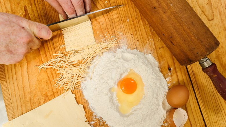 Close-up of hands cutting pasta dough, with flour, egg and rolling pin on a wooden table.