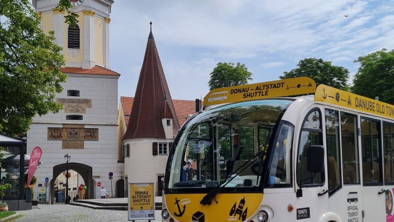 A white old town shuttle bus stands in front of a historic archway and a church tower.