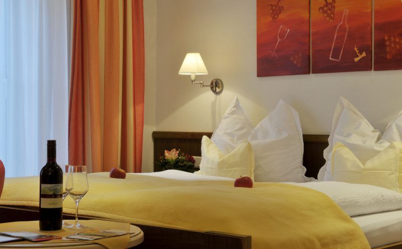 Gemütliches Hotelzimmer mit Doppelbett, gelber Bettdecke, Weinflasche und Gläsern auf Tisch, Wandkunst mit Weinmotiv.