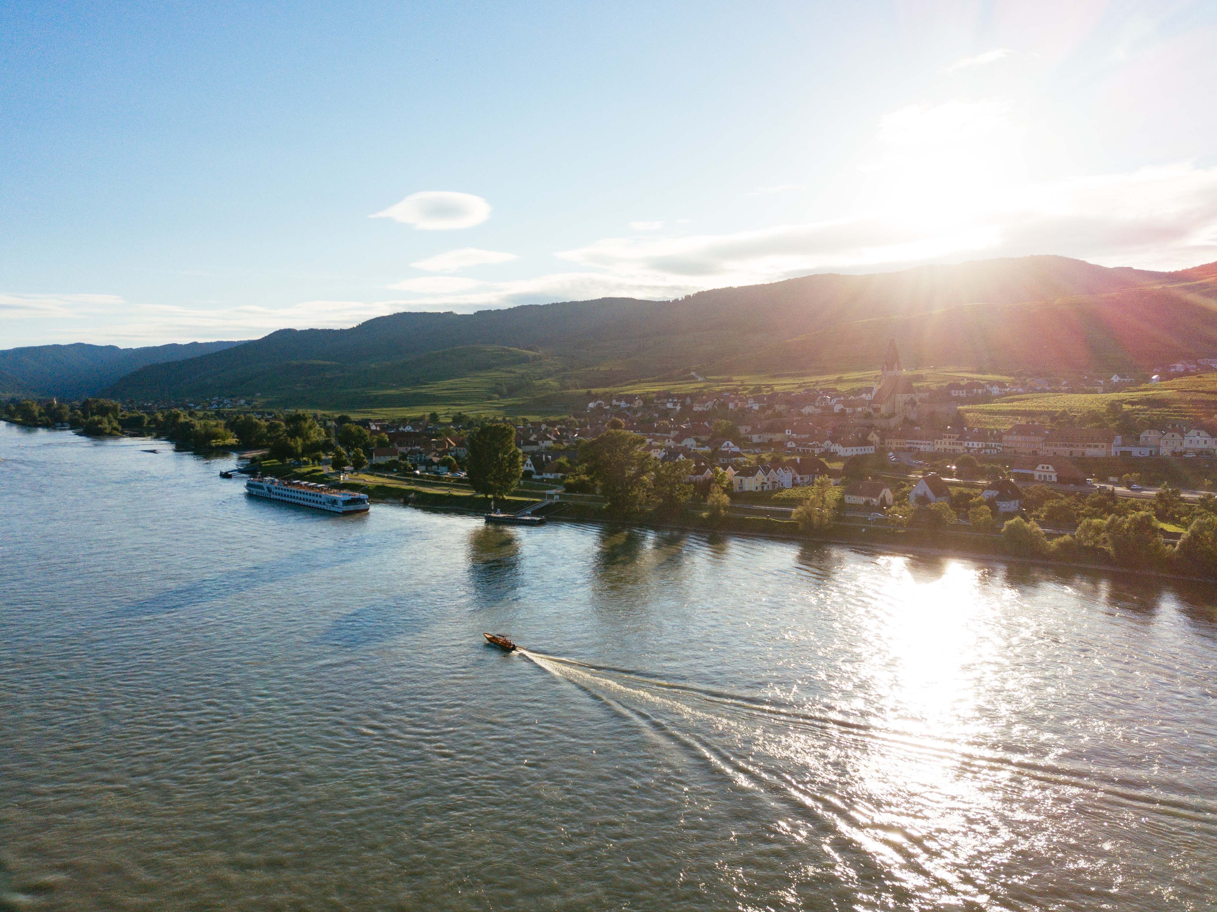 Luftaufnahme der Wachau mit Fluss, Booten und Dorf im Sonnenuntergang.