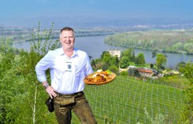 Ein Mann in traditioneller Kleidung h&auml;lt ein Tablett mit Essen vor einer Landschaft mit Fluss und Weinbergen.