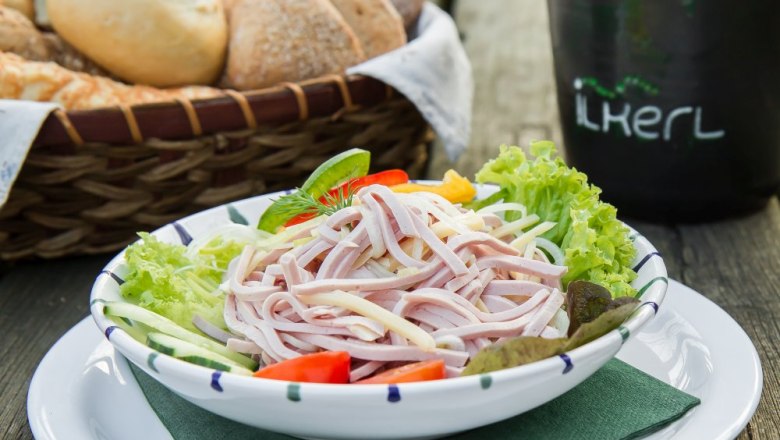Ein Teller mit Wurstsalat, garniert mit Salatbl&auml;ttern, Tomaten und Gurken. Im Hintergrund ein Brotkorb.