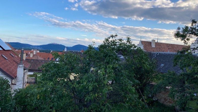 Blick &uuml;ber D&auml;cher und B&auml;ume auf H&uuml;gel und blauen Himmel mit Wolken.