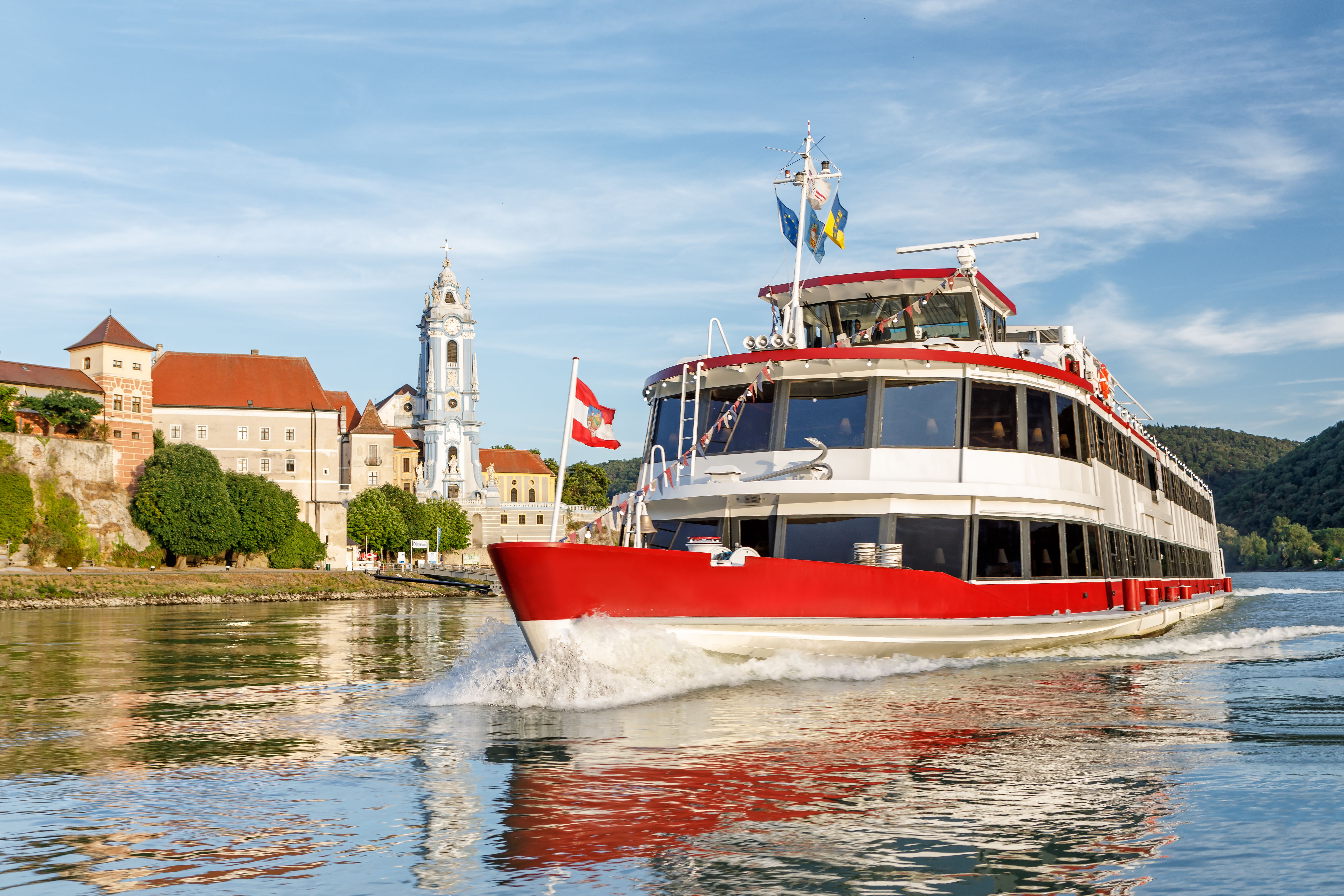 Ein rotes Passagierschiff fährt auf der Donau vor einer historischen Stadt mit einem markanten Kirchturm.