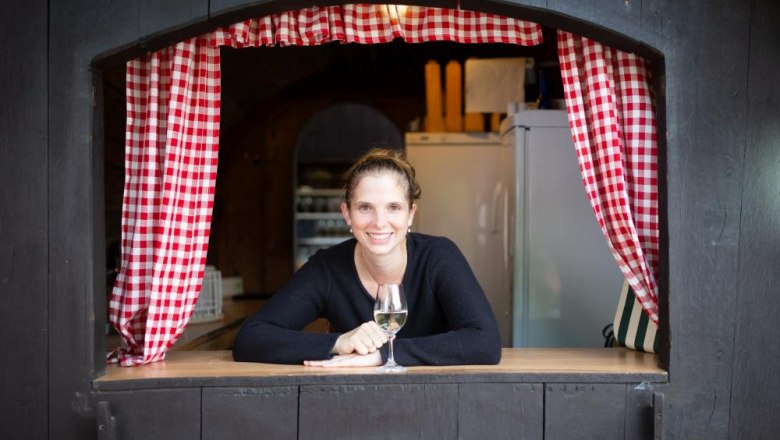 Woman leaning out of a window with red and white checkered curtains, holding a glass of wine.