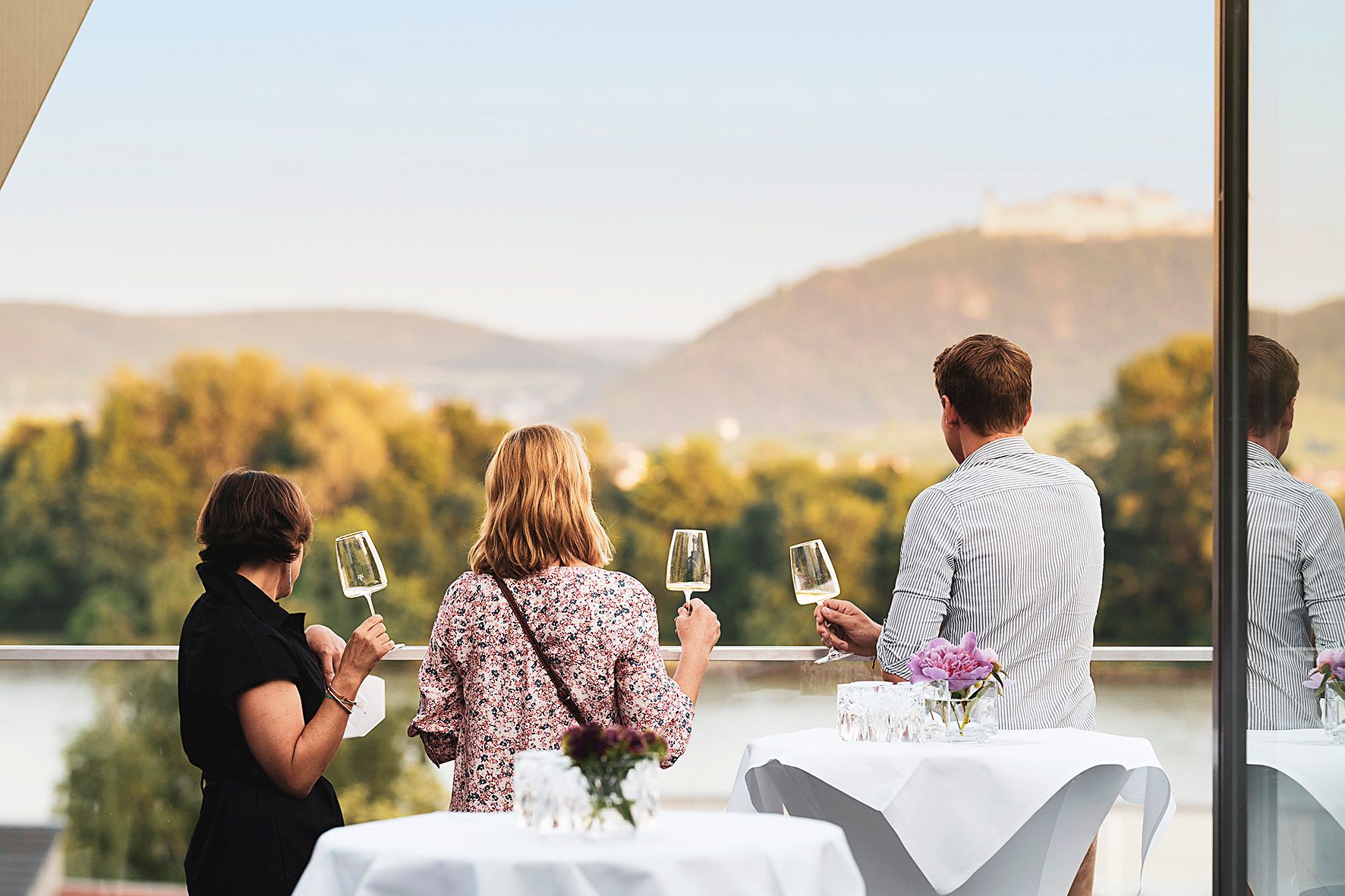 Drei Personen auf einer Terrasse mit Weingläsern, Blick auf Hügel und Fluss.