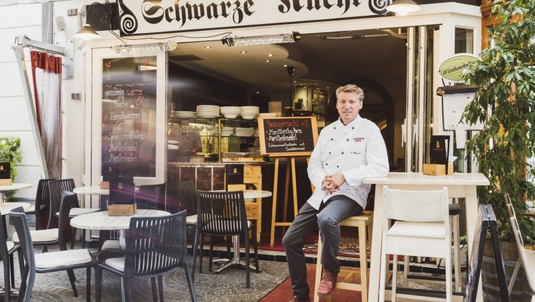 Restaurant 'Schwarze Kuchl' mit Koch vor dem Eingang, Tische im Freien.