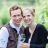 Ein l&auml;chelndes Paar in einem Weinberg, der Mann tr&auml;gt eine Weste, die Frau h&auml;lt ein Weinglas.