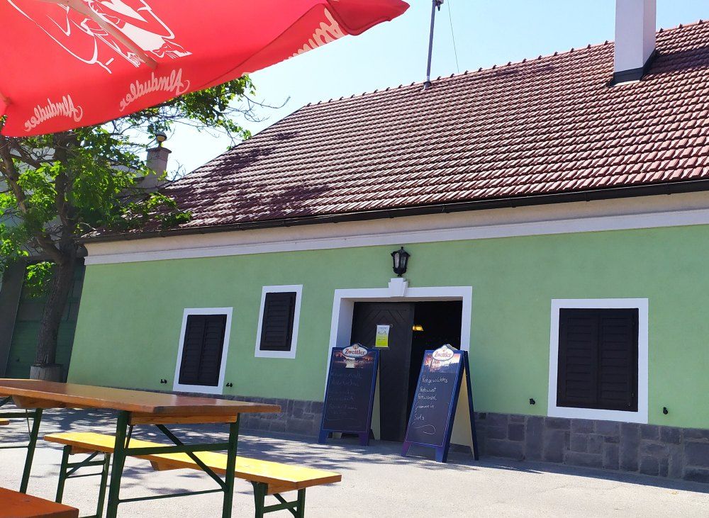 Außenansicht eines traditionellen Heurigen mit grünem Anstrich und roten Dachziegeln. Vor dem Gebäude stehen Holzbänke und ein roter Sonnenschirm.