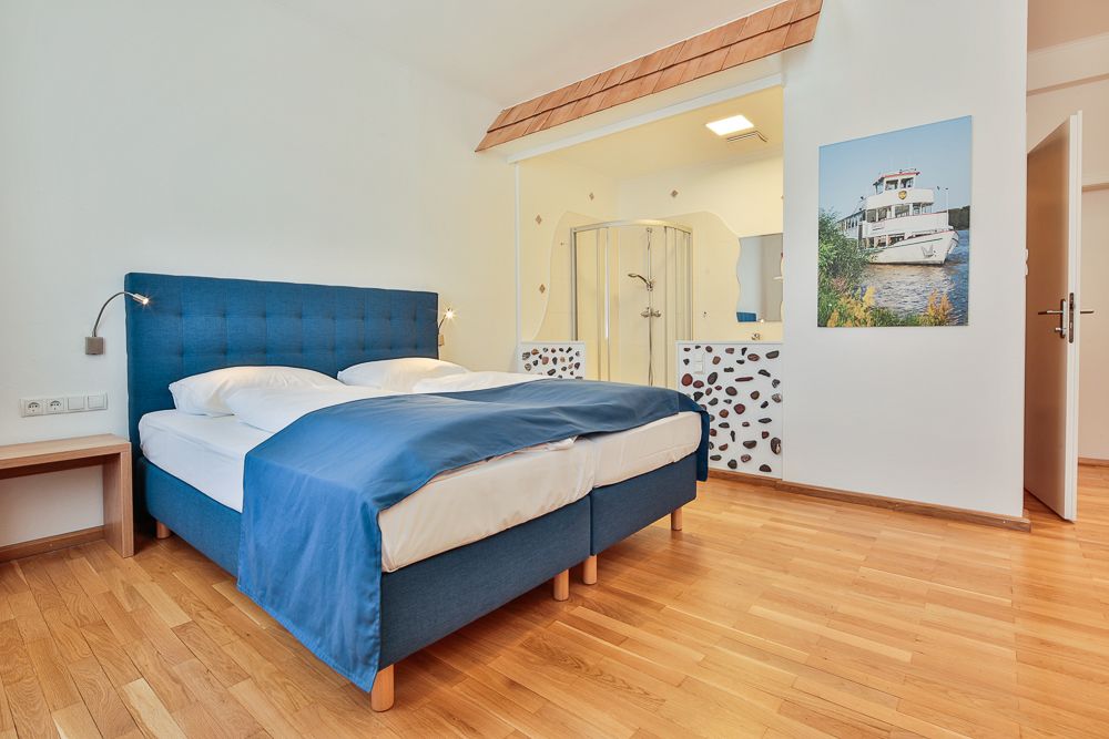 Hotelzimmer mit blauem Bett, Holzboden und offener Dusche.