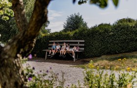 Four children sit on an oversized bench in a garden, surrounded by trees and bushes.