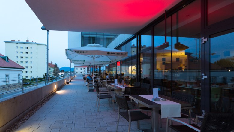 Terrace of a restaurant with tables and chairs, illuminated by red lights, with a view of buildings in the background.